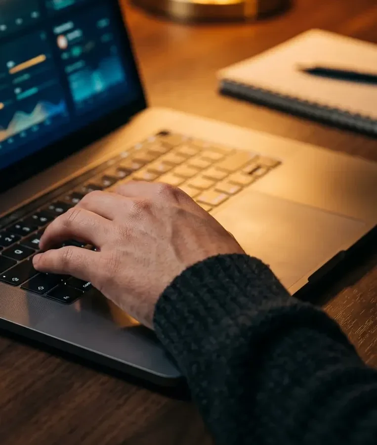 Detail of a hand on a laptop keyboard while a campaign report glows beside a warm brass lamp