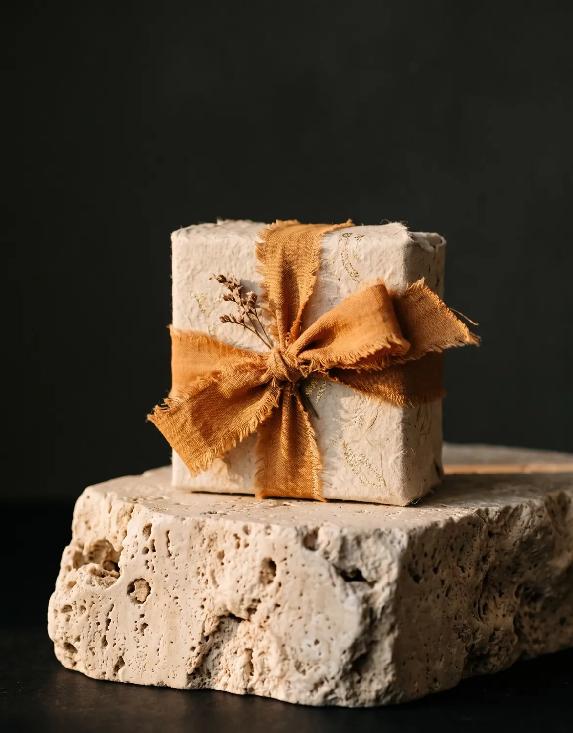 Gift box tied with an orange ribbon on a travertine stone block against a dark background, festive product advert