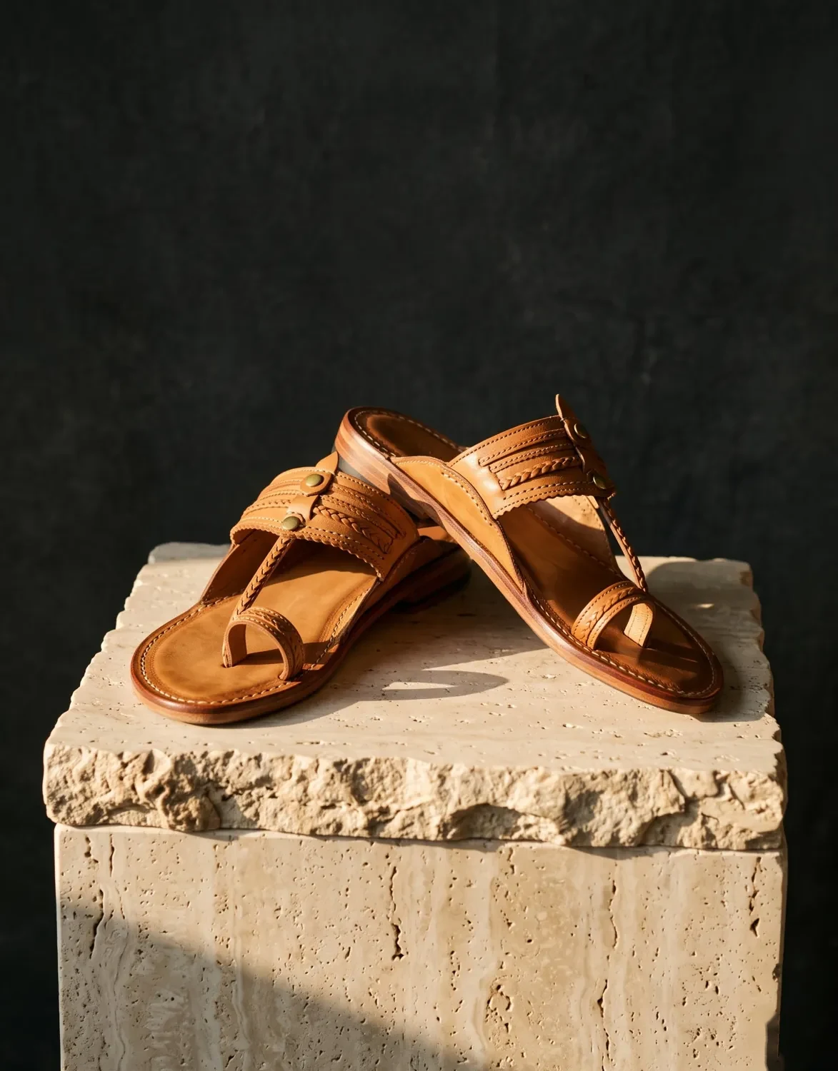 Pair of handcrafted leather sandals on a stone pedestal under warm side light on a dark backdrop, retail advert photo