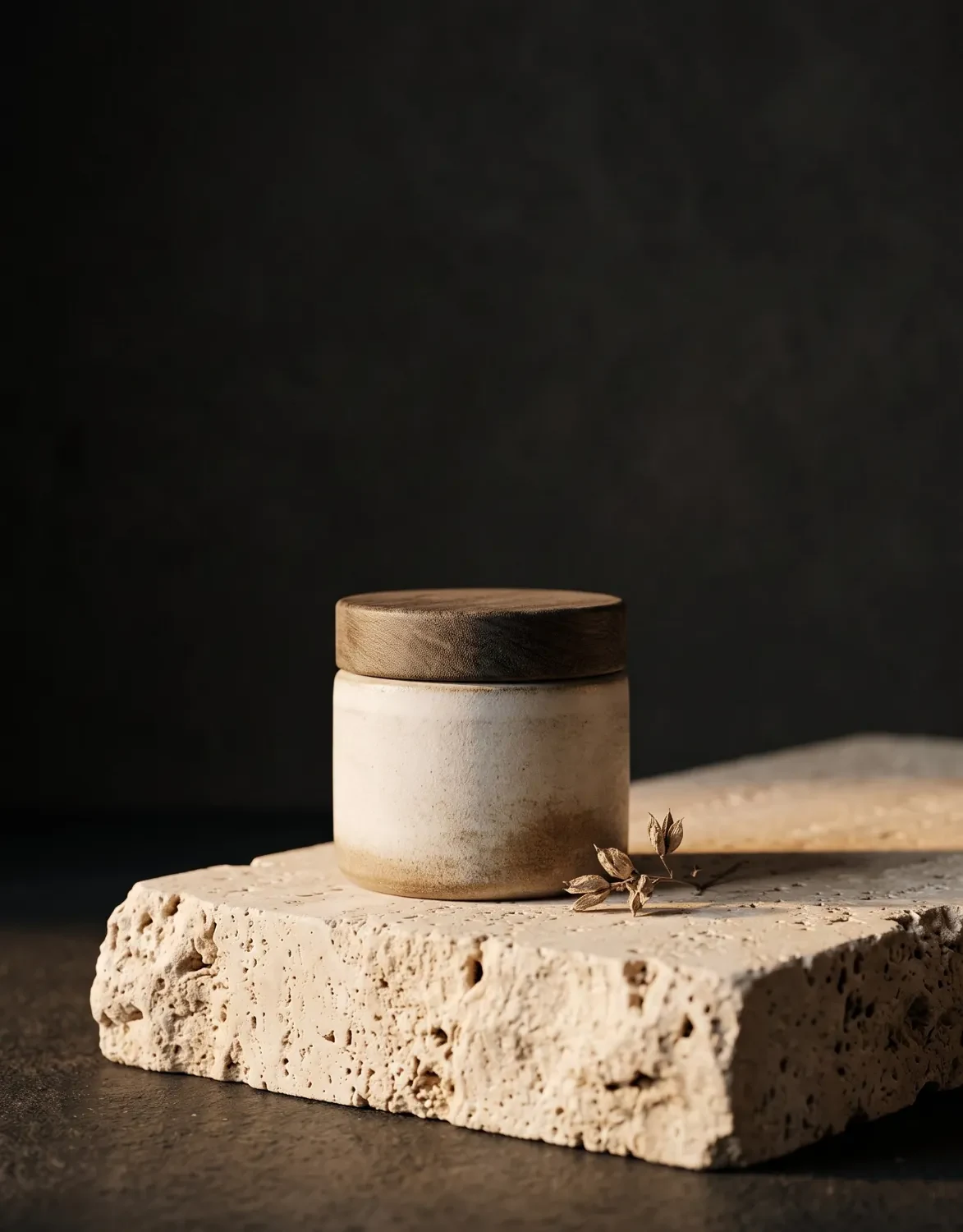 Ceramic skincare jar on a rough travertine pedestal with soft side light on a dark backdrop, premium product advert